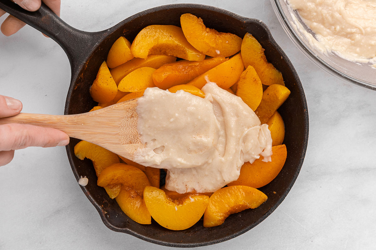 Το Cobbler Topping που εξαπλώνεται πάνω από φρέσκα ροδάκινα σε μια κατσαρόλα από χυτοσίδηρο.